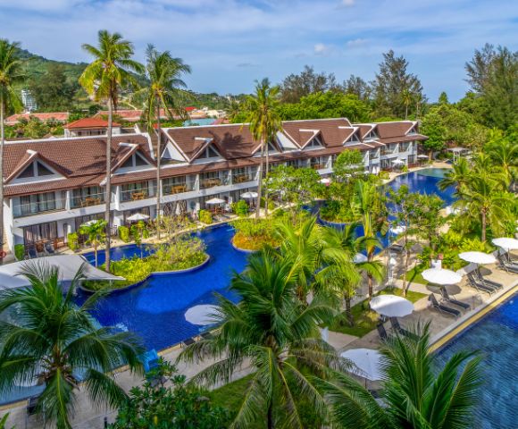 Tropical resort with a series of bungalows beside a winding blue pool, surrounded by palm trees and lush greenery. Lounge chairs and umbrellas line the water.