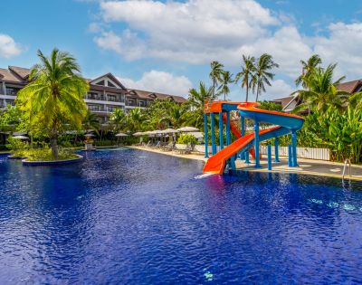 Tropical resort pool with bright red and blue waterslide, surrounded by palm trees and sun loungers, under a partly cloudy sky. Calm, inviting atmosphere.