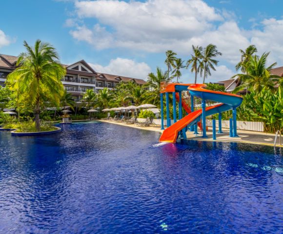 Tropical resort pool with bright red and blue waterslide, surrounded by palm trees and sun loungers, under a partly cloudy sky. Calm, inviting atmosphere.