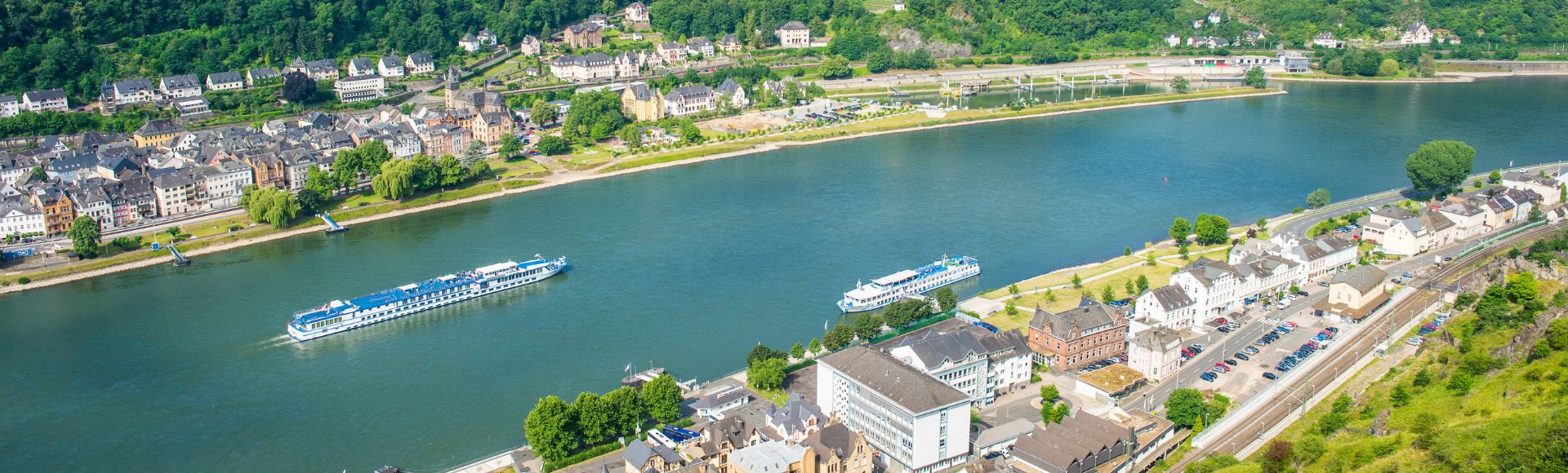 Aerial view of a scenic river, with two cruise ships gliding along. Lush hills and quaint town buildings line the river, creating a tranquil, picturesque scene.