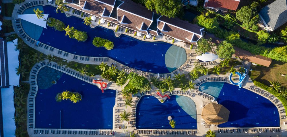 Aerial view of a large, curvy swimming pool complex surrounded by lounge chairs and palm trees. The scene is serene and vibrant, exuding a tropical resort vibe.