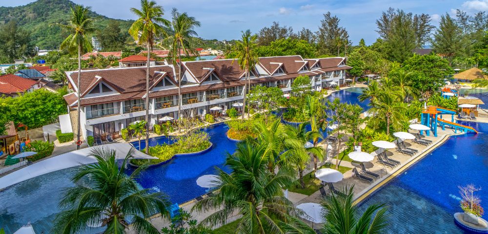 Tropical resort with a series of bungalows beside a winding blue pool, surrounded by palm trees and lush greenery. Lounge chairs and umbrellas line the water.