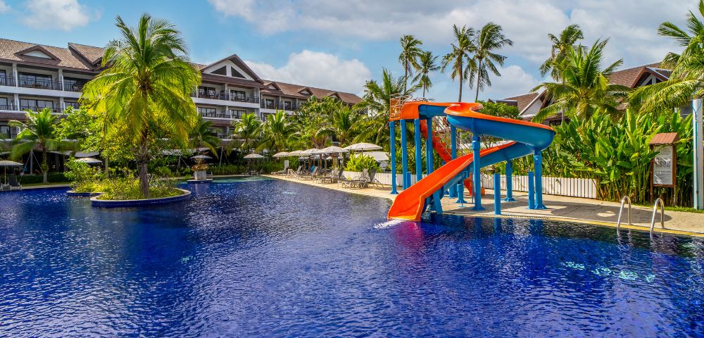 Tropical resort pool with bright red and blue waterslide, surrounded by palm trees and sun loungers, under a partly cloudy sky. Calm, inviting atmosphere.