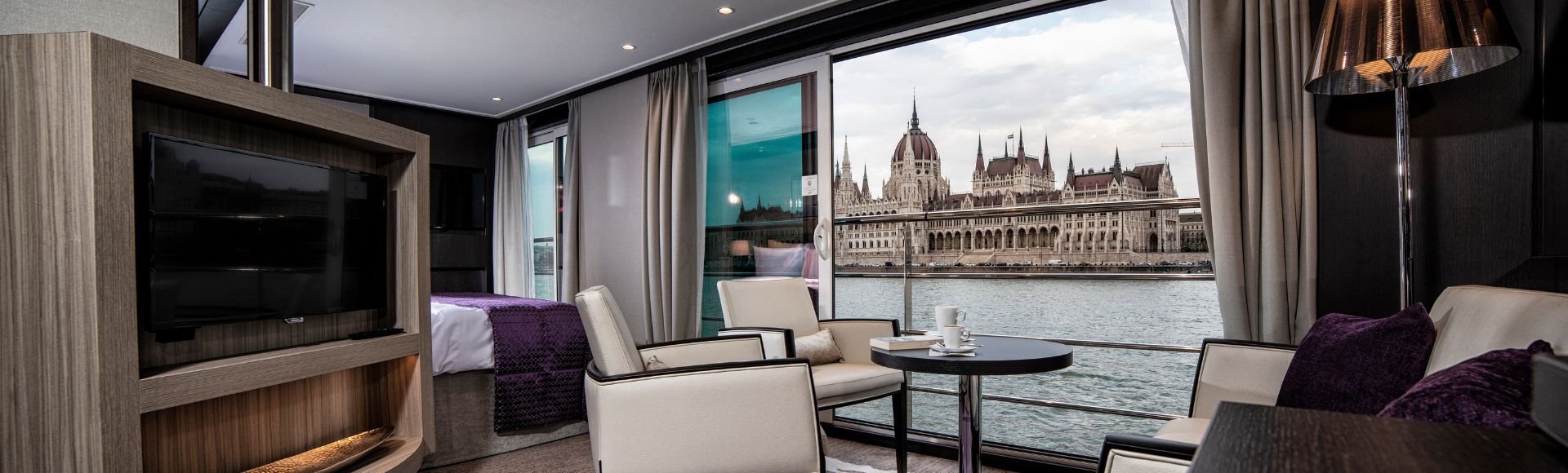 Luxurious riverboat suite with modern white chairs, purple accents, and a view of a grand historic building across the river through large windows.
