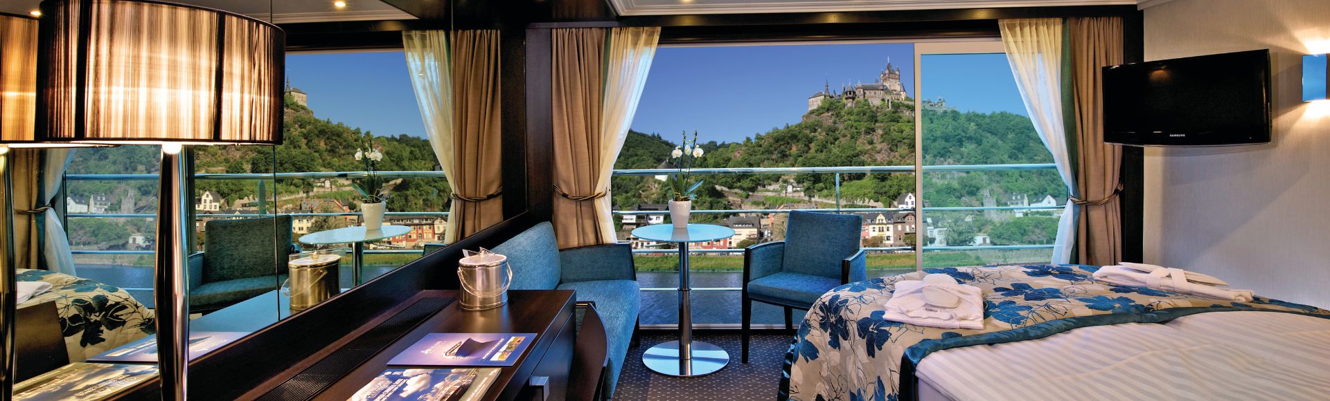 Luxurious cruise ship cabin with large window view of a hilltop castle. Room features a cozy bed, sitting area, elegant decor, and a calming ambiance.