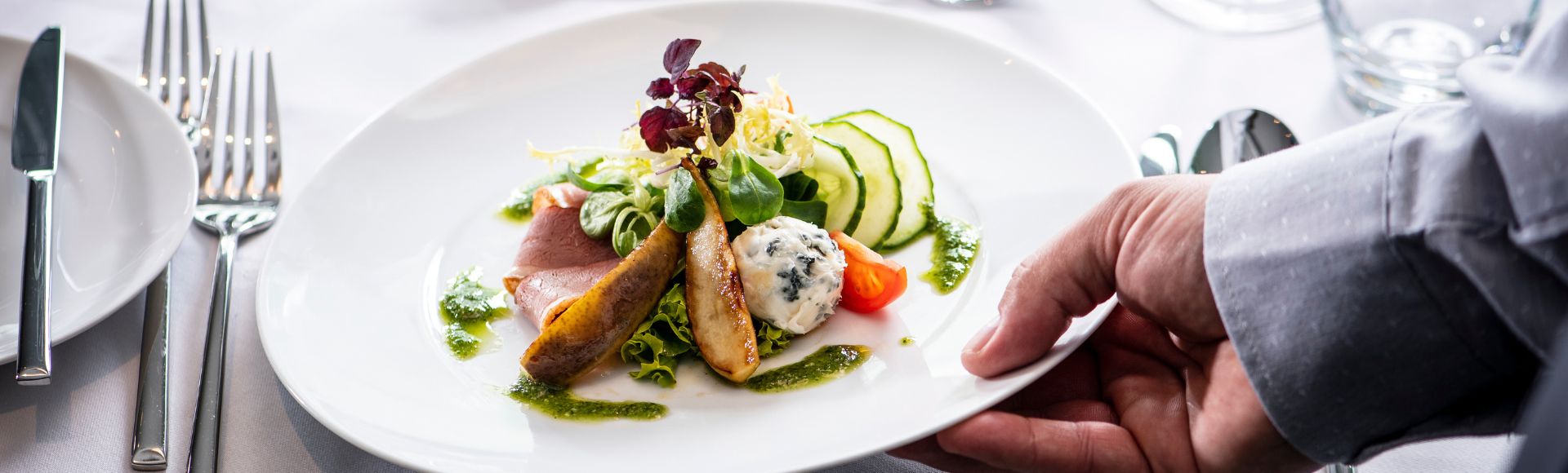 A hand serves a gourmet dish on a white plate featuring colorful vegetables, sliced cucumber, and artful garnishes on a white linen table setting.