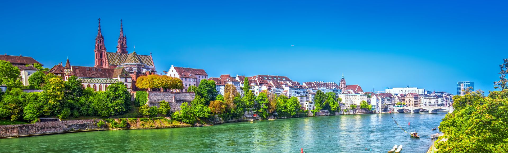 A scenic view of Basel, Switzerland, featuring the historic Münster Cathedral with its twin spires, surrounded by lush greenery along the Rhine River.