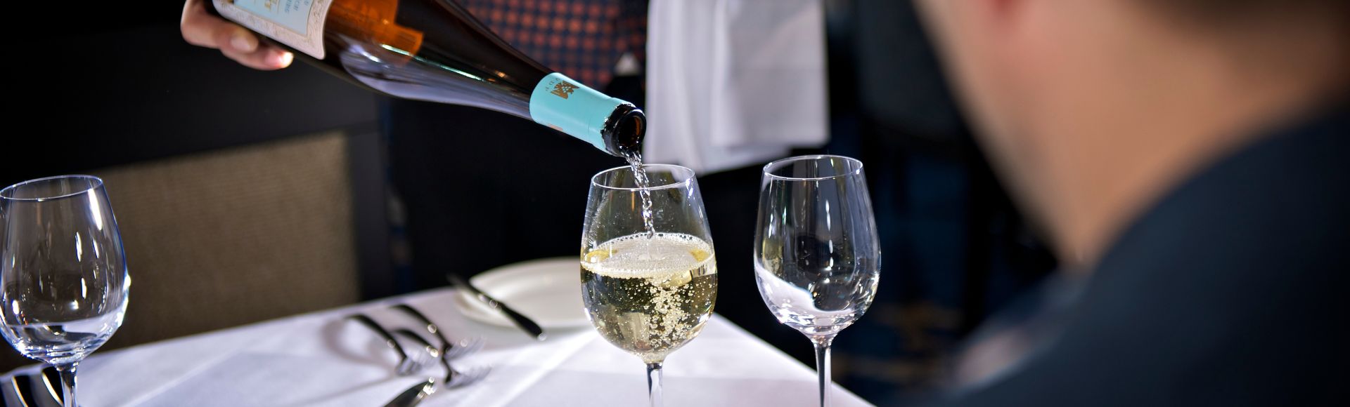 A person pours white wine into a glass at a table set with cutlery and empty glasses. The scene conveys a sense of elegance and anticipation.