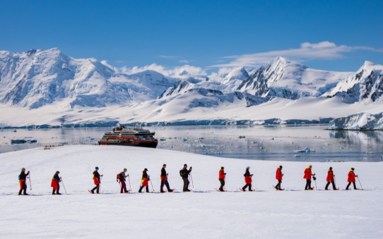 A group of people in red jackets hike in a line across a snow-covered landscape. A large ship floats on calm icy waters beneath snow-capped mountains.