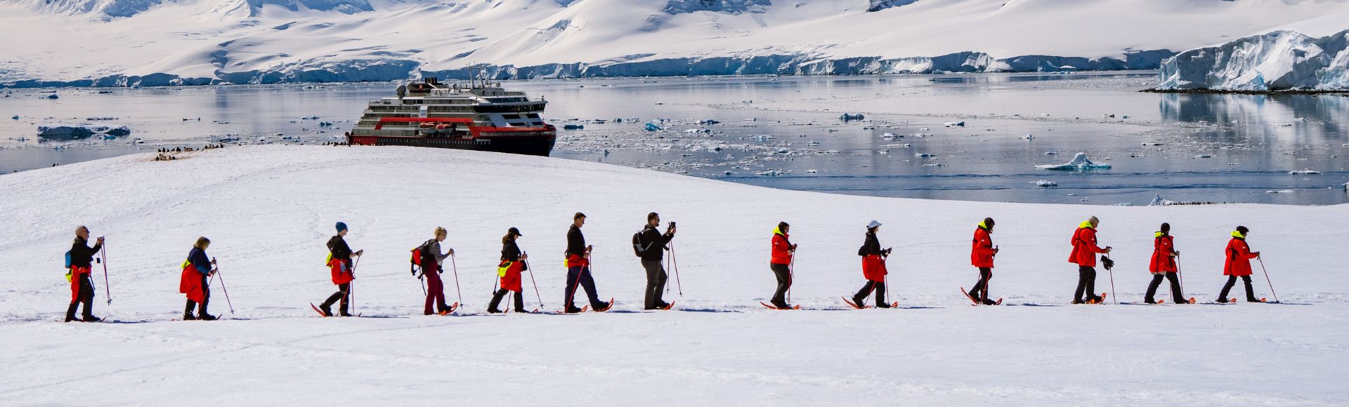 A group of people in red jackets hike in a line across a snow-covered landscape. A large ship floats on calm icy waters beneath snow-capped mountains.