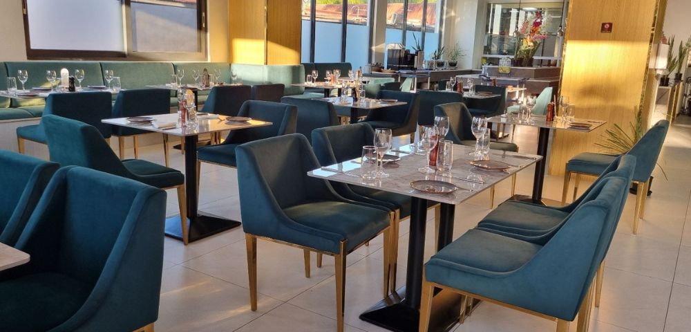 Modern restaurant interior with teal velvet chairs and marble tables set with glassware and cutlery. Soft, warm lighting creates a cozy atmosphere.