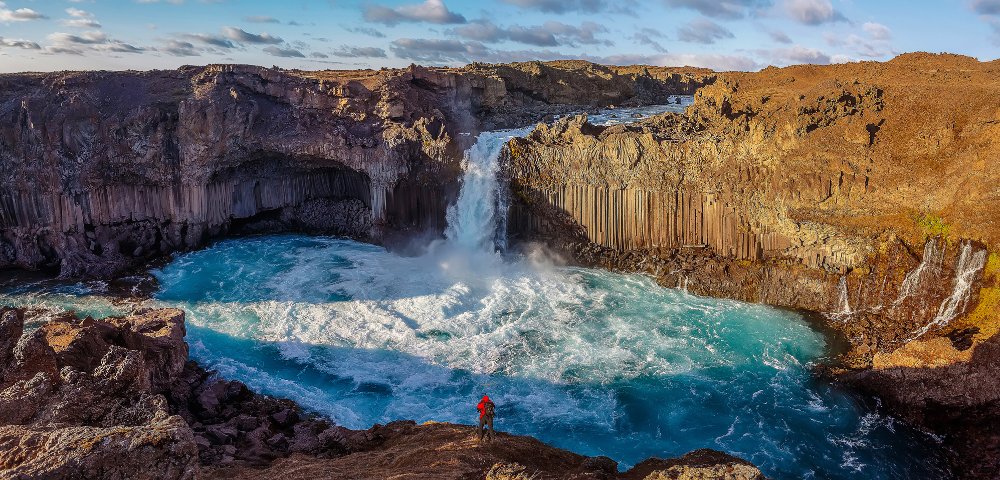 A person in a red jacket stands before a vibrant blue waterfall in a rocky canyon under a partly cloudy sky, evoking a sense of awe and adventure.