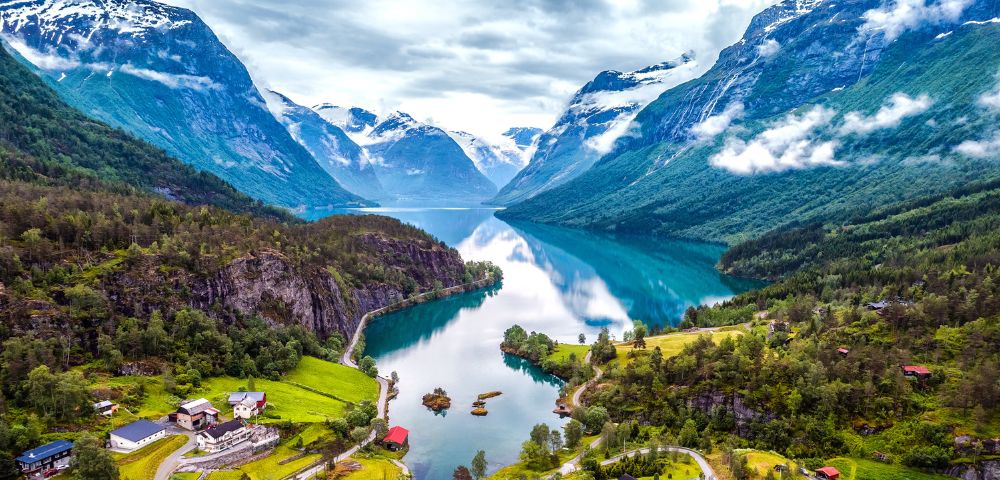 A serene fjord landscape with emerald waters, surrounded by snow-capped mountains and lush green forests. Cozy houses dot the shoreline, evoking tranquility.