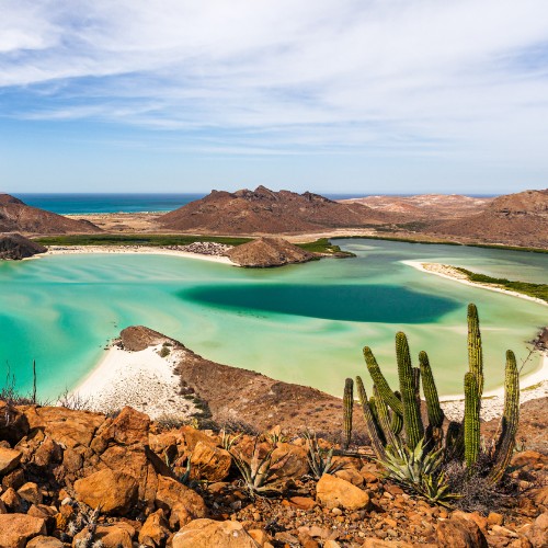 Aerial view of a tranquil bay with turquoise waters, surrounded by rocky hills and cacti under a blue sky with wispy clouds, conveying a serene desert seascape.