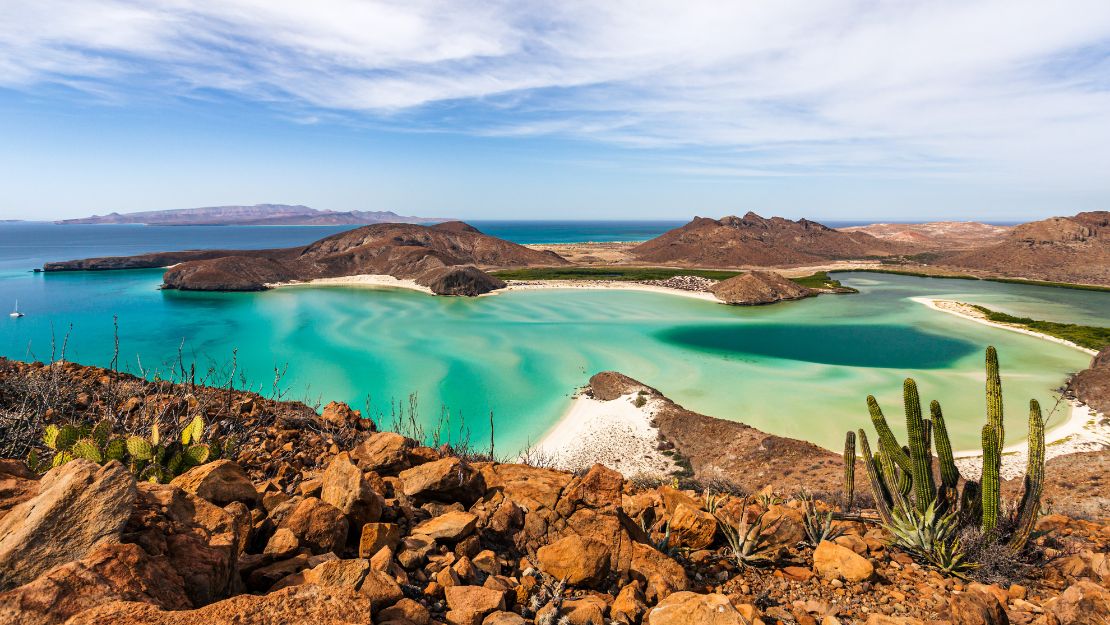 Aerial view of a tranquil bay with turquoise waters, surrounded by rocky hills and cacti under a blue sky with wispy clouds, conveying a serene desert seascape.