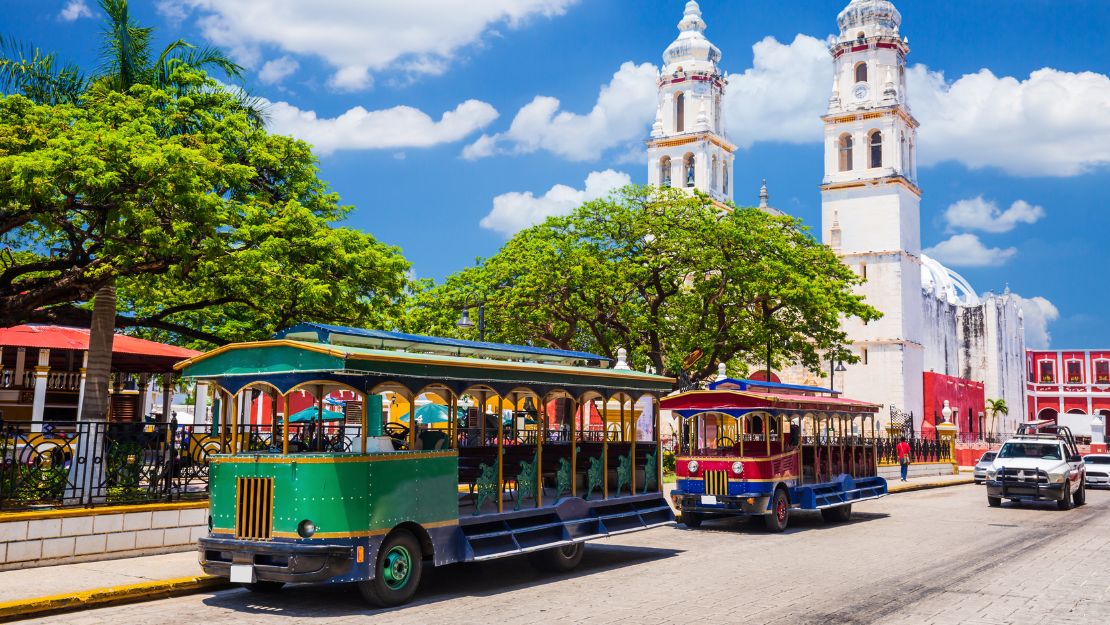 Colorful open-air tourist trams drive by a historic church with two tall towers. Lush green trees and a bright blue sky create a vibrant scene.