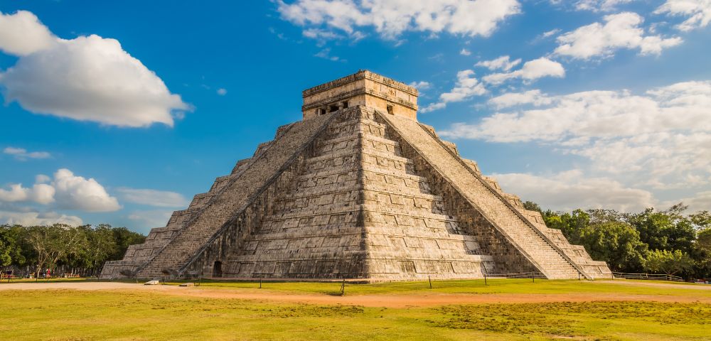 Ancient stone pyramid of Chichen Itza stands under a vibrant blue sky with scattered clouds, surrounded by lush green grass, evoking awe and history.