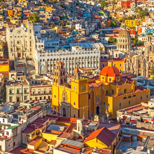 A vibrant aerial view of Guanajuato, featuring colorful buildings densely packed along hilly terrain. Prominent landmarks include a yellow church and ornate cathedral.