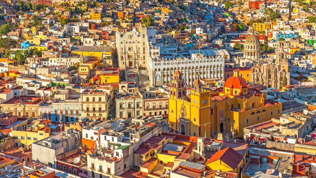 A vibrant aerial view of Guanajuato, featuring colorful buildings densely packed along hilly terrain. Prominent landmarks include a yellow church and ornate cathedral.