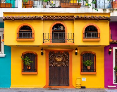 Bright yellow and orange building facade with ornate wooden doors. Arched windows have wrought iron balconies and potted plants, evoking a warm, inviting tone.