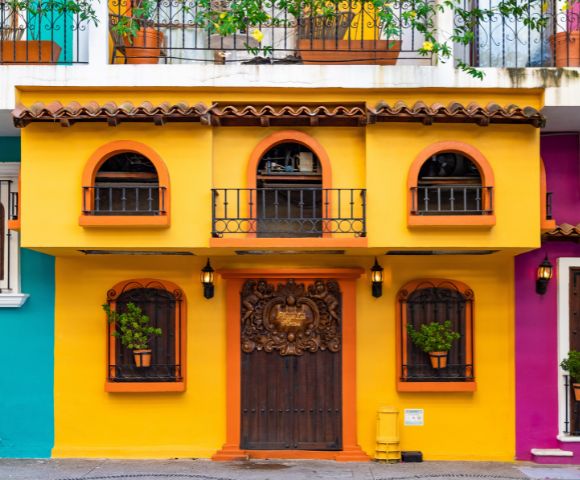 Bright yellow and orange building facade with ornate wooden doors. Arched windows have wrought iron balconies and potted plants, evoking a warm, inviting tone.