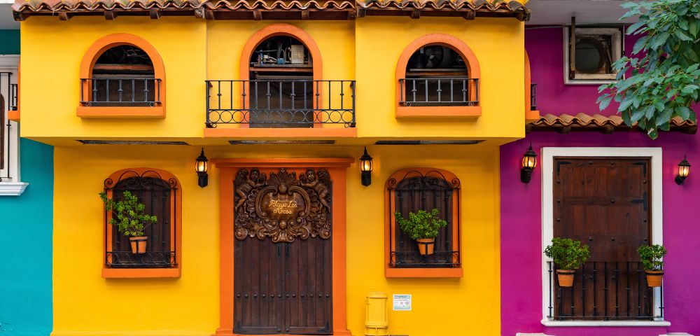 Bright yellow and orange building facade with ornate wooden doors. Arched windows have wrought iron balconies and potted plants, evoking a warm, inviting tone.
