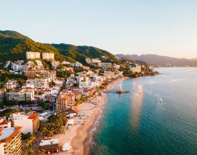 Coastal cityscape at sunset with lush hills, vibrant buildings, and a tranquil beach lining the turquoise waters. A serene, warm atmosphere prevails.