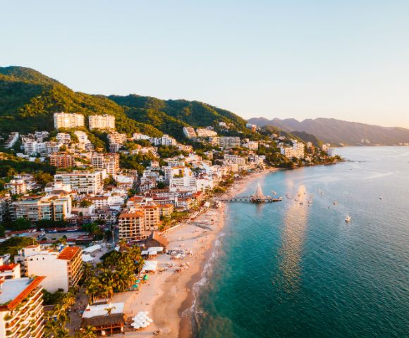 Coastal cityscape at sunset with lush hills, vibrant buildings, and a tranquil beach lining the turquoise waters. A serene, warm atmosphere prevails.