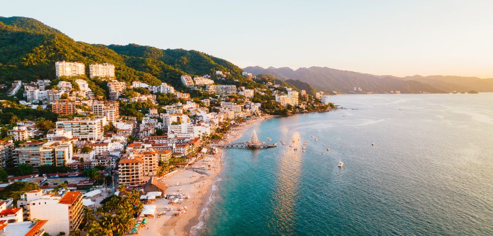Coastal cityscape at sunset with lush hills, vibrant buildings, and a tranquil beach lining the turquoise waters. A serene, warm atmosphere prevails.