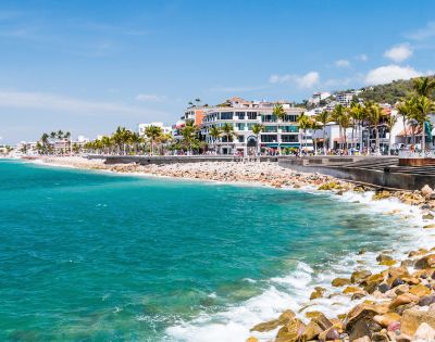 Coastal cityscape with turquoise ocean waves meeting a rocky shore. Palm-lined promenade with colorful buildings under a bright blue sky. Vibrant and lively.