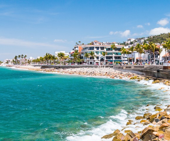Coastal cityscape with turquoise ocean waves meeting a rocky shore. Palm-lined promenade with colorful buildings under a bright blue sky. Vibrant and lively.