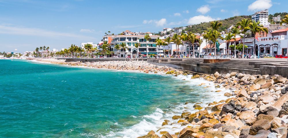 Coastal cityscape with turquoise ocean waves meeting a rocky shore. Palm-lined promenade with colorful buildings under a bright blue sky. Vibrant and lively.
