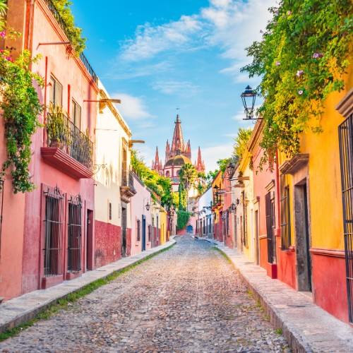 Cobblestone street in San Miguel de Allende with colorful buildings adorned with greenery, leading to a distant, ornate church under a bright blue sky.