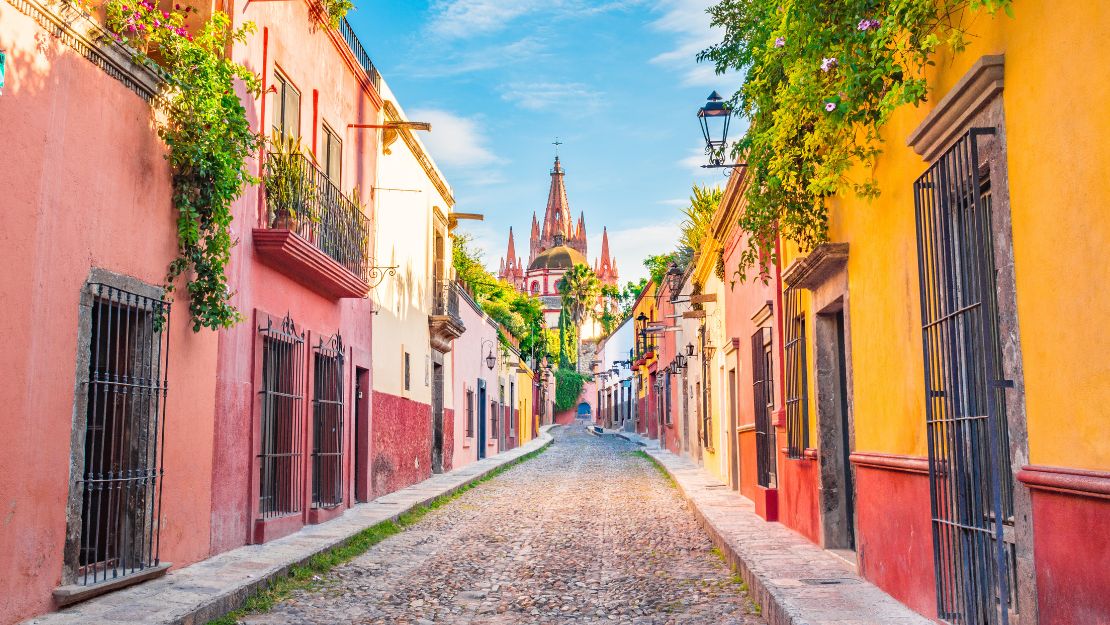 Cobblestone street in San Miguel de Allende with colorful buildings adorned with greenery, leading to a distant, ornate church under a bright blue sky.