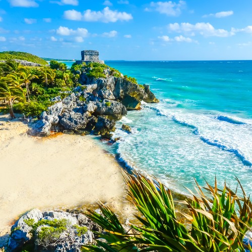 Sunny beach scene at Tulum, Mexico featuring ancient ruins on a cliff. Clear turquoise waves lap against the sandy shore with lush greenery nearby.