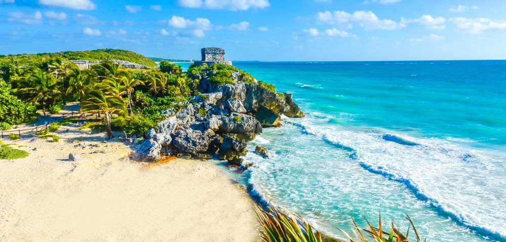 Sunny beach scene at Tulum, Mexico featuring ancient ruins on a cliff. Clear turquoise waves lap against the sandy shore with lush greenery nearby.