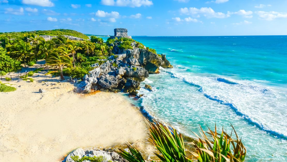 Sunny beach scene at Tulum, Mexico featuring ancient ruins on a cliff. Clear turquoise waves lap against the sandy shore with lush greenery nearby.