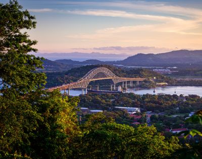 Scenic view of a large arch bridge spanning a river, surrounded by lush greenery and mountains under a pastel sunset sky, conveying tranquility.