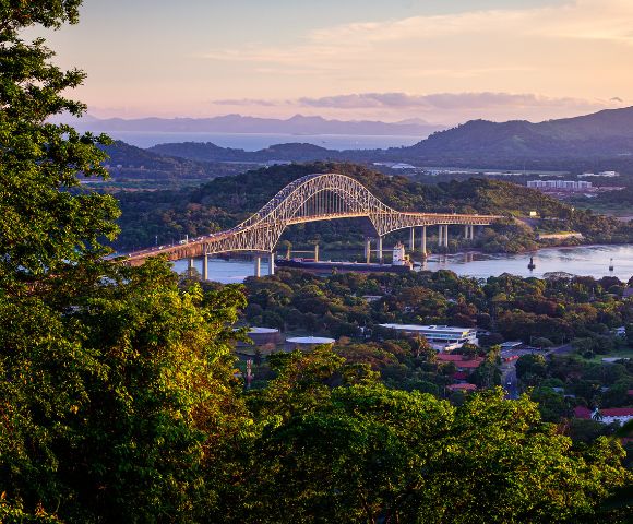 Scenic view of a large arch bridge spanning a river, surrounded by lush greenery and mountains under a pastel sunset sky, conveying tranquility.