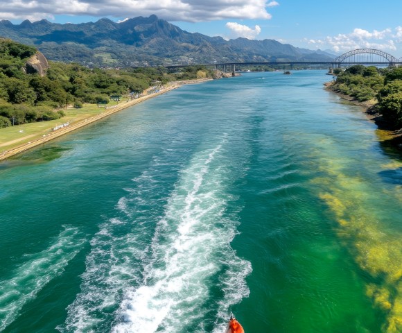 A boat glides along a vibrant green river under a clear blue sky with fluffy clouds, flanked by lush greenery and distant mountains. Serene and scenic.