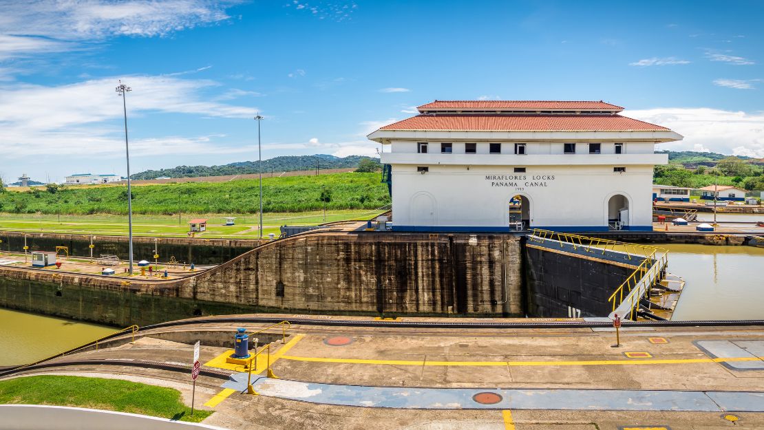 Panama Canal's Miraflores Locks on a sunny day, featuring the control building with a red-tiled roof, surrounded by greenery and blue skies.