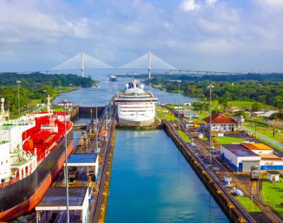 Aerial view of the Panama Canal with a cruise ship passing through the lock. Surrounding greenery and a distant bridge under a blue sky enhance the scene.