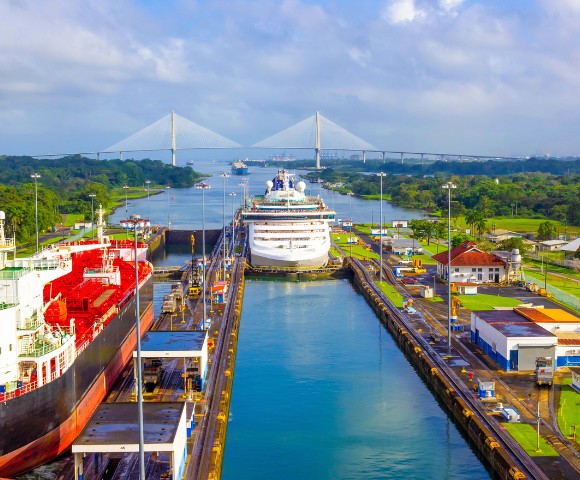 Aerial view of the Panama Canal with a cruise ship passing through the lock. Surrounding greenery and a distant bridge under a blue sky enhance the scene.