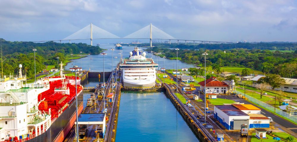 Aerial view of the Panama Canal with a cruise ship passing through the lock. Surrounding greenery and a distant bridge under a blue sky enhance the scene.
