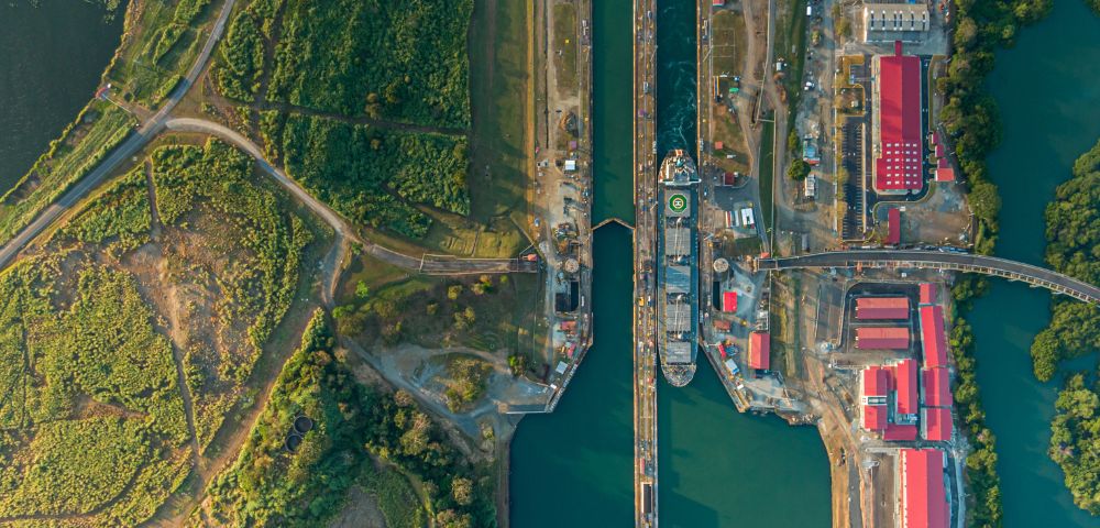 Aerial view of the Panama Canal with a ship passing through a lock. Lush greenery surrounds the canal, with red-roofed buildings on either side.