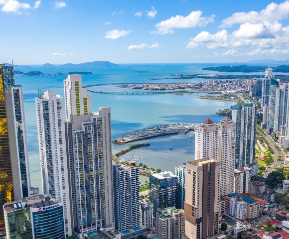 Aerial view of a coastal cityscape with tall skyscrapers, a waterfront, and a distant bridge. Blue ocean under a clear sky with scattered clouds.