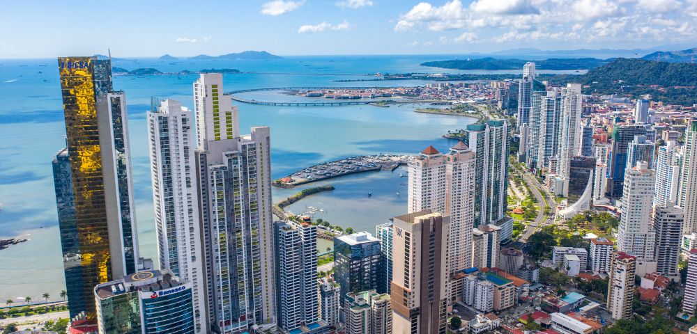 Aerial view of a coastal cityscape with tall skyscrapers, a waterfront, and a distant bridge. Blue ocean under a clear sky with scattered clouds.