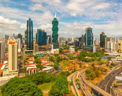 A vibrant cityscape featuring a mix of modern skyscrapers and greenery under a partly cloudy sky. Busy roads curve through the bustling urban landscape.