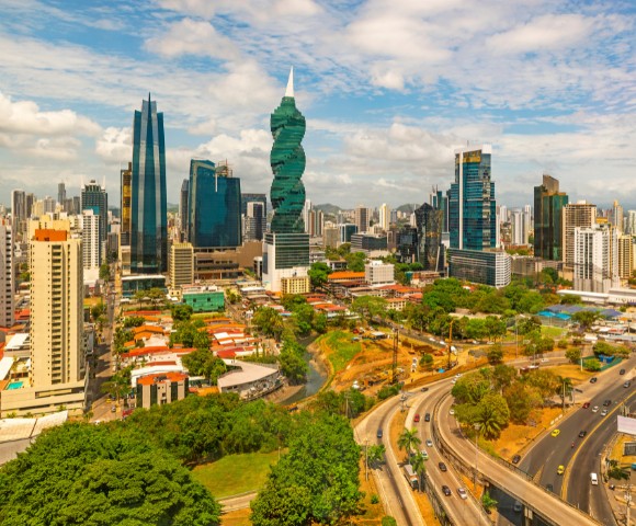 A vibrant cityscape featuring a mix of modern skyscrapers and greenery under a partly cloudy sky. Busy roads curve through the bustling urban landscape.