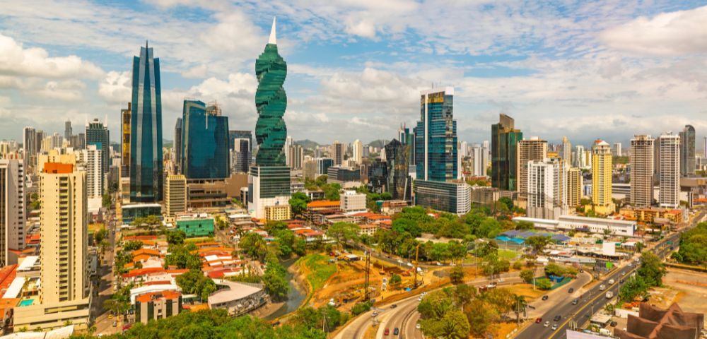 A vibrant cityscape featuring a mix of modern skyscrapers and greenery under a partly cloudy sky. Busy roads curve through the bustling urban landscape.